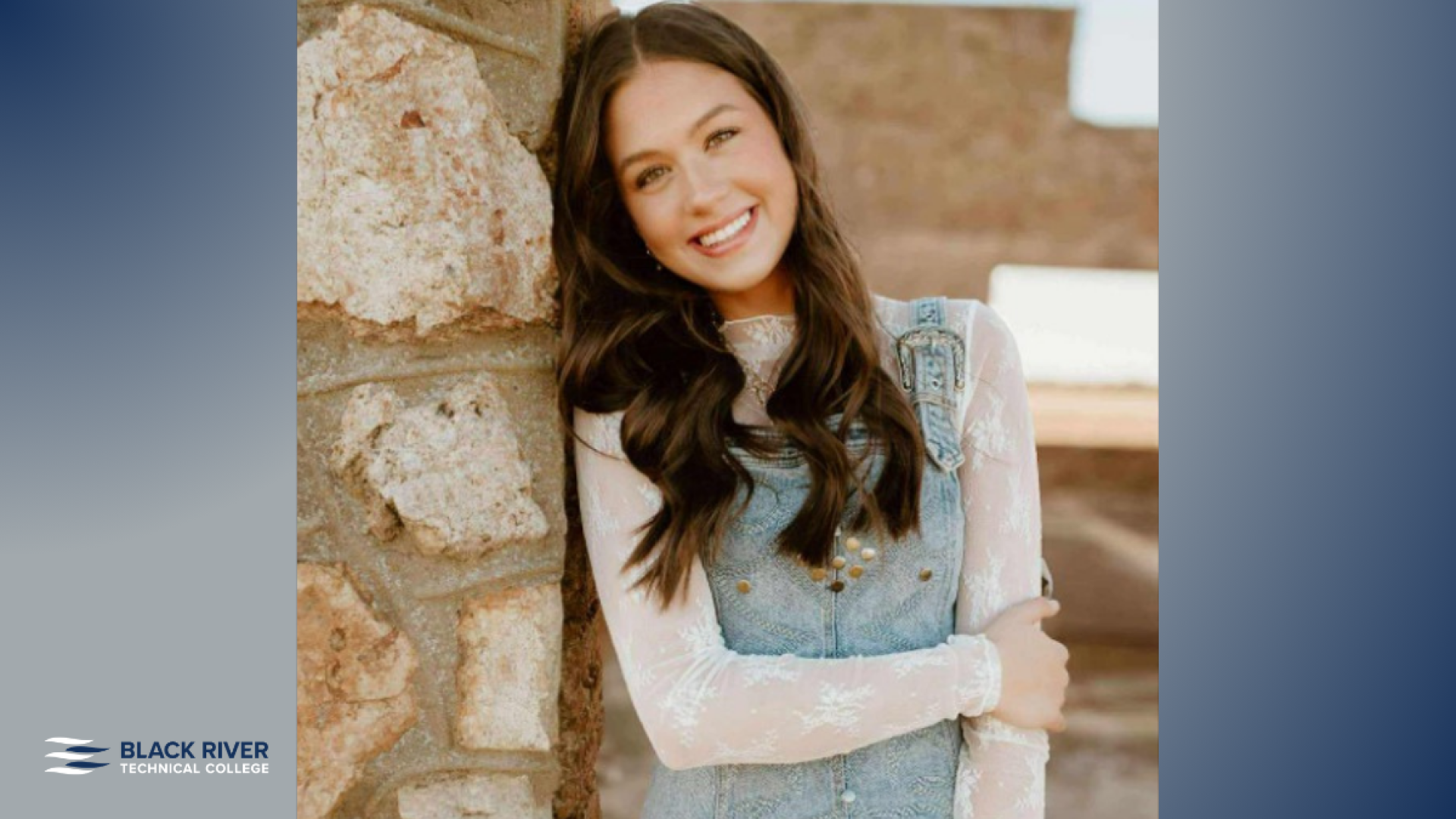 A young woman with long dark wavy hair, wearing denim overalls over a white lace top, smiles while leaning against a stone wall, with Black River Technical College branding.